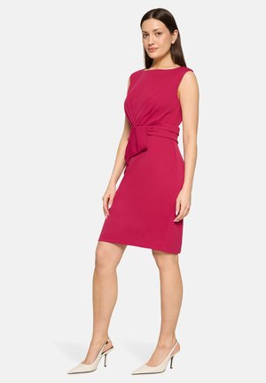 Kvinde, der står i en ærmeløs knælængde magenta kjole med sløjfedetalje i siden, iført hvide slingback høje hæle mod en ensfarvet baggrund.