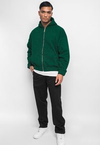 Grønn hettegenser med glidelås foran, store lommer og ribbestrikkede mansjetter. Brukt sammen med en hvit skjorte og svarte bukser, komplementert med hvite sneakers.