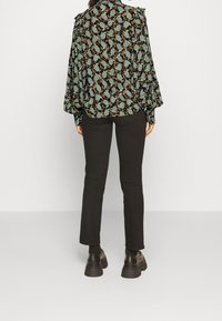Blusa nera a maniche lunghe con motivo di foglie verdi e marroni, abbinata a pantaloni neri aderenti e scarpe nere chunky. Vista posteriore.