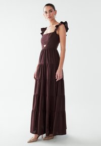 Maxi-jurk in kastanjebruin met gelaagd ontwerp, vierkante halslijn en gerimpelde mouwen. Gemaakt van lichtgewicht stof, met een uitgesneden detail aan de taille.