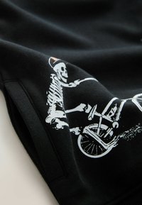 Un squelette blanc portant une casquette fait du vélo, imprimé sur un tissu noir, montrant une partie de la manche et le détail de la couture.