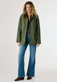 Giacca esterna verde con colletto marrone, chiusure a pressione, tasche laterali, indossata sopra una camicia a righe e jeans svasati azzurri chiari, abbinata a stivali marroni.