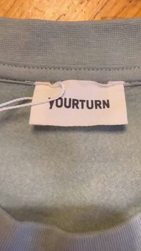 Pelēks kapuces krekls ar adījuma faktūru, ar balto birku, kurā melnā krāsā ir rakstīts "YOURTURN". Birka piestiprināta ar aukliņu.