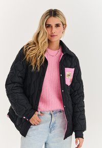 Žena s dlouhými blond vlasy, oblečená v černé prošívané bundě přes růžový svetr a světle modré džíny, stojící před jednobarevným bílým pozadím.