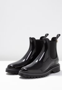 Botas de tobillo impermeables negras, hechas de material brillante, con paneles laterales elásticos, lengüetas para tirar y suela de goma texturada.