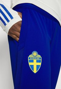 Modré sportovní kalhoty s texturovanou tkaninou, na boku s žluto-modrým fotbalovým logem Švédska a postranní kapsou. Viditelný bílý rukáv trička.