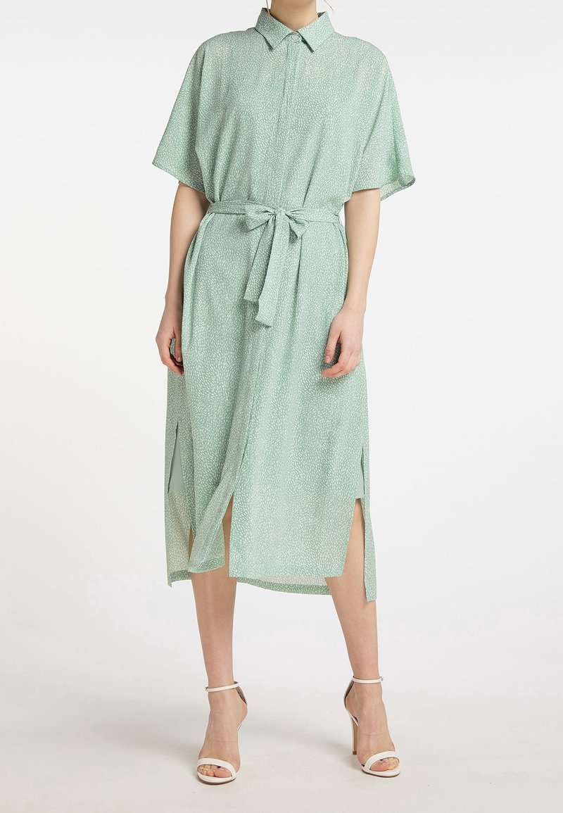 Femme portant une robe midi vert clair à manches courtes, boutonnée, avec une ceinture à la taille et des fentes hautes sur les côtés, associée à des sandales blanches à talons hauts.