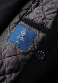 Doublure en tissu matelassé noir avec une étiquette bleue portant la mention "OMBRE HERITAGE." Comprend un détail de bouton transparent et un motif en losanges dans le tissu.