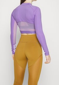Fialový krátký top s dlouhými rukávy a průsvitným zadním dílem, kombinovaný s hnědými legínami s vysokým pasem, zdobenými šikmými výřezy a švovými detaily.