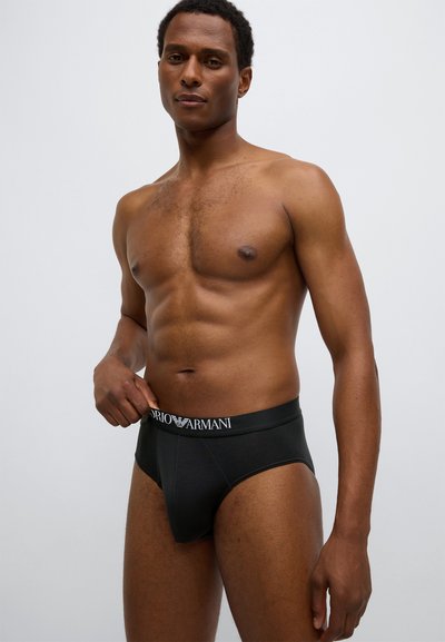 Mustat bokserit, joissa on valkoinen logo sisältävä merkitty vyötärönauha. Kangas näyttää sileältä ja istuu tiiviisti vartalolle, korostaen muotoa.