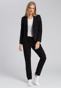 Schwarzer maßgeschneiderter Blazer mit passender Hose, kombiniert mit einem weißen Oberteil und weißen Sneakers. Glatter Stoff, figurschnittige Passform und minimalistische Knöpfe.