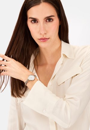 Montre en argent avec un cadran rond et un bracelet à maillons, portée au poignet. La personne qui la porte est vêtue d'une blouse légère à col.