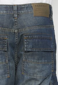 Sötétkékszínű farmer cargo nadrág, texturált felülettel, számos oldalsó zsebbel és egy strapabíró bőrlogóval a derékrészen.