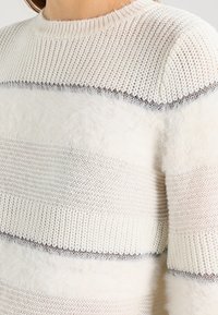 Vit och krämfärgad randig tröja med stickade och fluffiga texturer, har rund halsringning och en kontrasterande grå ränder.