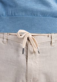 Jasnoniebieski sweter dziany z prążkowanym dołem, noszony z kremowymi, przewiewnymi spodniami, które mają związany sznurek z metalowymi końcówkami.