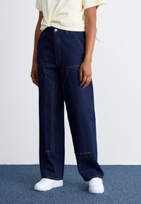 Calças de ganga de cintura alta em azul-escuro com detalhes de costura em destaque, corte largo e bolsos frontais, combinadas com ténis brancos e um top amarelo-claro.