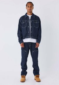 Conjunto de chaqueta y pantalones de denim en azul oscuro con patrón de diamantes blancos. La chaqueta tiene bolsillos y botones. Botas marrones completan el atuendo.