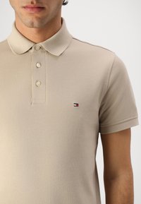 Muškarac u bež polo majici kratkih rukava s tri gumba i malim crveno-bijelo-plavim logotipom na prsima.