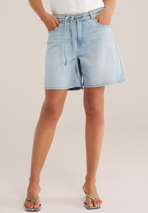 Világoskék farmer rövidnadrág kötött övvel, fehér felsővel és kígyómintás szandállal viselve, kezét zsebbe téve, semleges háttér előtt állva.