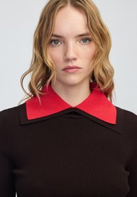 Młoda kobieta z falującymi blond włosami, ubrana w ciemny prążkowany top z szerokim czerwonym kołnierzem, patrzy bezpośrednio w kamerę z neutralnym wyrazem twarzy.