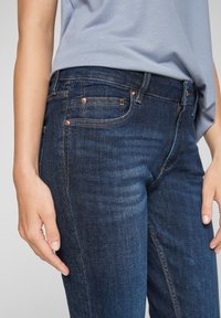 Dark blue denim jeans with a fitted design, accentuated stitching, copper hardware, and five pocket style. Texture appears smooth and slightly worn.