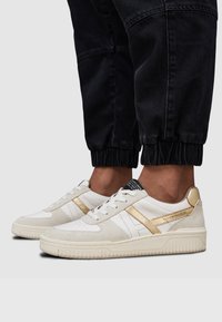 Białe i beżowe sneakersy z złotymi akcentami, wykonane z zamszu i skóry, z teksturowanymi gumowymi podeszwami i perforowanymi panelami bocznymi.