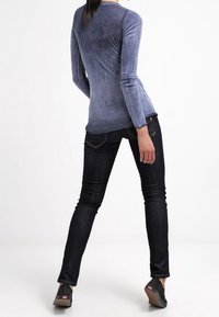 Top ajustado de manga larga en color verde azulado con una textura suave, combinado con jeans ajustados oscuros. Los zapatos son negros con cuña baja y un detalle sutil del logo.