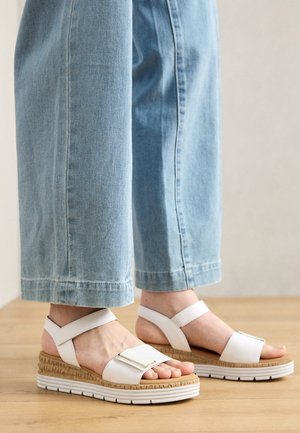 Witte sandalen met een dikke espadrille-achtige zool, voorzien van een glimmende rechthoekige gespsluiting en enkelsandalen, gecombineerd met lichtblauwe wijde spijkerbroeken.