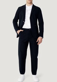 Σετ με μπλε μπλέιζερ του ναυτικού και ταιριαστό παντελόνι από λείο ύφασμα, με εφαρμοστή γραμμή. Φοριέται με λευκό πουκάμισο και λευκά αθλητικά παπούτσια.