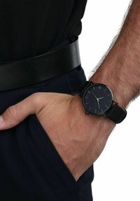 Schwarze Uhr mit minimalistischem Design, ausgestattet mit einem runden Ziffernblatt, silbernen Markierungen und einem schwarzen Lederarmband, das auf einer Hand ruht.