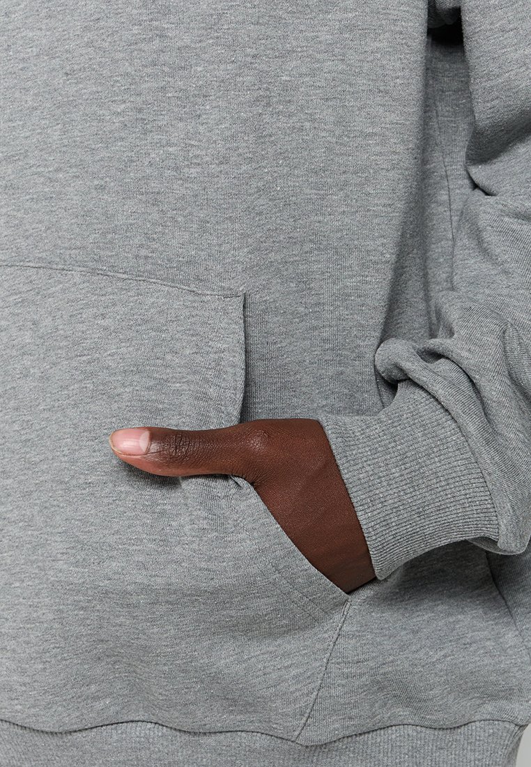 Mano de piel oscura colocada dentro del bolsillo frontal de un sudadera gris claro con puños acanalados en la manga.