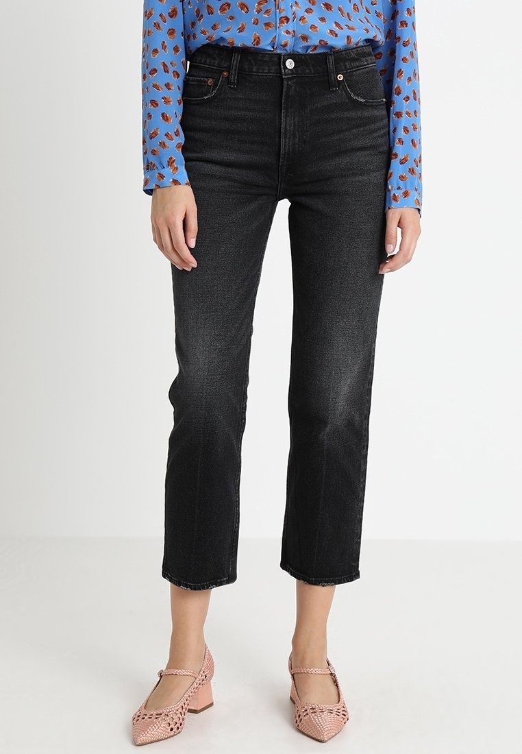 Jeans neri a vita alta con gamba dritta, dotati di chiusura con bottone e cinque tasche, abbinati a una blusa blu a fantasia e scarpe rosa.