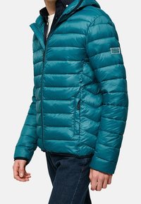 Teal puffjacka med huva, som har vertikala kviltade paneler, fickor med dragkedja och en subtil logotyp på överarmen.