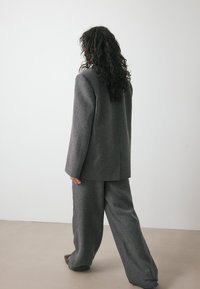 Hallhalli tekstuuriga bleiser ja sobivad laiad püksid, millel on täpselt mõõdetud lõige ja pikad varrukad, nähtavad tagant neutraalses taustas.