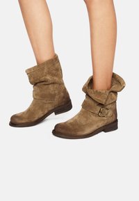 Botas de tobillo de ante marrón con un borde enrollado en la parte superior, correa con una hebilla de metal y detalles texturizados en el borde. Tacón bajo de madera apilada.