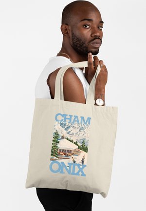 Uomo con maglietta bianca senza maniche che tiene una borsa a tracolla beige con illustrazione di un rifugio di montagna e sciatori etichettata "Cham Onix."