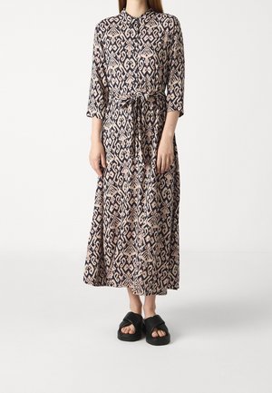 Woman wearing a long-sleeve, patterned midi dress with a front tie belt and black slip-on platform sandals, standing on a plain white background.