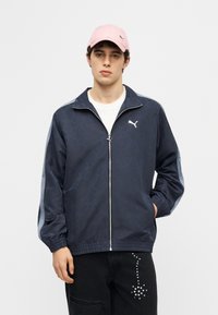 Νεαρός άνδρας που φοράει ζακέτα Puma σε ναυτικό μπλε με φερμουάρ, λευκό μπλουζάκι, μαύρο παντελόνι με σχέδιο από τρουκς και ένα ανοιχτό ροζ καπέλο Puma, στέκεται μπροστά σε ένα απλό φόντο.