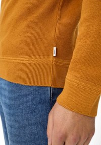 Orangefarbener Strickpullover mit gerippten Ärmeln und Saum. Verfügt über ein kleines Etikett an der Seite. Trägt man mit blauen Jeans. Die Textur ist gewoben.