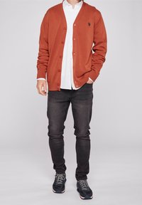 Rust-colored cardigan with a ribbed texture, button front, and embroidered logo. Worn over a white shirt and black jeans with dark sneakers.