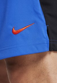Blå sportshorts med en röd Nike-swoosh-logotyp som bärs på ett naket ben, bredvid ett svart nätmaterial.