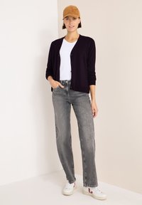 Dunkel lila Cardigan, weißes T-Shirt, graue Jeans und weiße Sneaker. Eine braune Kappe akzentuiert das Outfit. Glatte Texturen und lockere Passform.