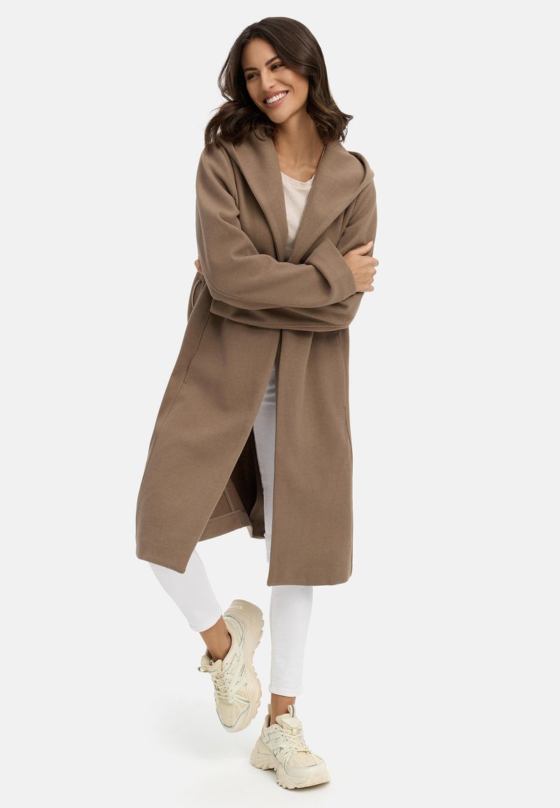 Long manteau marron avec une large capuche, à l'avant ouvert et avec des poches latérales, fabriqué en tissu doux, associé à un pantalon blanc et des baskets beiges.