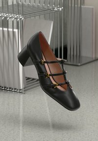 Black leather heeled shoe with square toe, featuring decorative straps and gold buckles. Set against a minimalist backdrop.