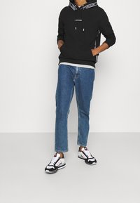 Felpa nera con cappuccio e logo, orlo inferiore grigio a strati, abbinata a jeans blu e sneakers nere/bianche con dettagli rossi.