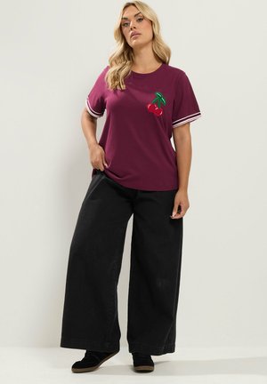 Vrouw draagt een paars T-shirt met kersenpatch en gestreepte mouwen, zwarte wijde broek en zwarte schoenen, staand tegen een effen achtergrond.