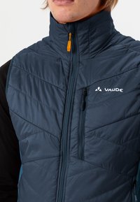 Gilet isolato blu navy con colletto alto, trapuntatura a zigzag, chiusura frontale con zip e tasca sul petto. Presenta il logo Vaude e un tirazip arancione.