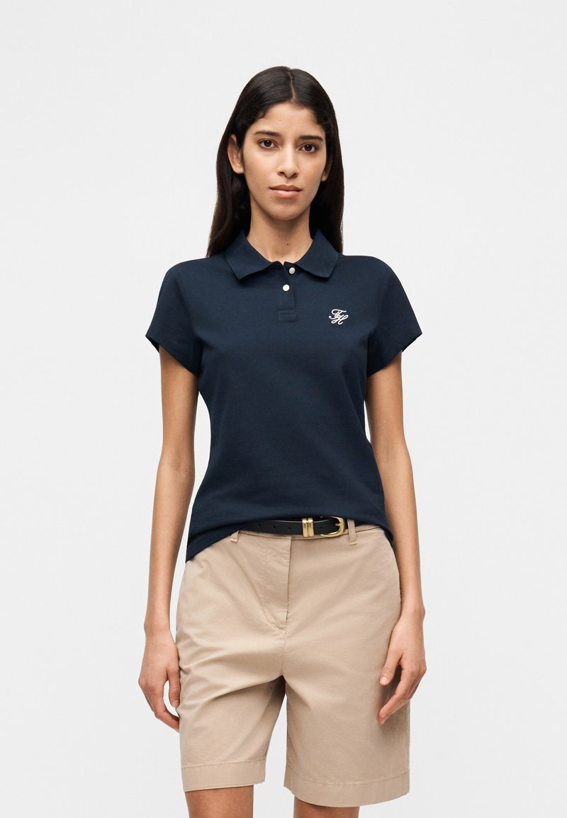 Γυναίκα που φοράει μπλε-σκούρο πόλο μπλουζάκι με κοντά μανίκια και λογότυπο, καθώς και μπεζ σορτς μήκους μέχρι το γόνατο με μαύρη ζώνη, στέκεται μπροστά σε απλό φόντο.