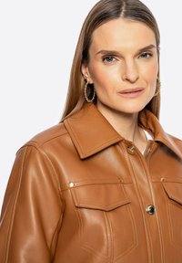 Giacca di pelle marrone con colletto, due tasche sul petto e dettagli in metallo dorato. La modella indossa orecchini a cerchio.