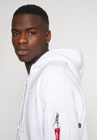 Sweat à capuche blanc avec cordon de serrage ajustable, poche latérale zippée, logo discret sur la poitrine et tissu texturé. Gros plan du modèle.